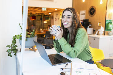Lächelnde Geschäftsfrau mit Kaffee in der Nähe von Laptop im Büro - WPEF08416