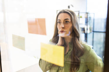 Thoughtful businesswoman examining adhesive notes on glass wall in office - WPEF08431