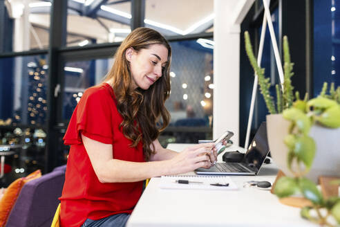Lächelnde reife Geschäftsfrau, die ein Smartphone am Schreibtisch im Büro benutzt - WPEF08457