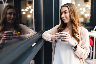 Lächelnde Geschäftsfrau mit Kaffeebecher am Fenster im Büro - WPEF08471