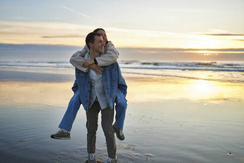 Smiling boyfriend piggybacking girlfriend on beach at sunset - ANNF00889