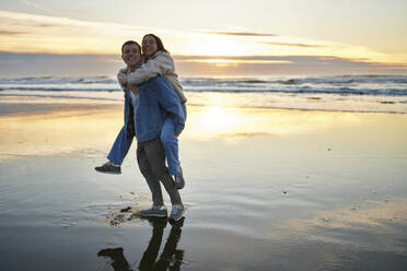 Smiling man carrying girlfriend piggyback at ocean beach - ANNF00890