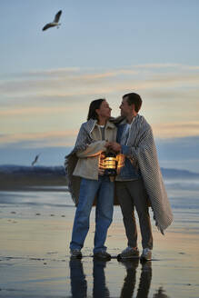 Lächelnder Mann und Frau mit Laterne, eingewickelt in ein Tuch, am Meeresstrand - ANNF00897