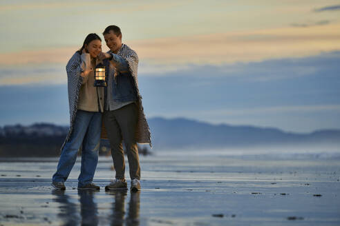 Lächelndes Paar mit Laterne am Strand stehend bei Sonnenuntergang - ANNF00899
