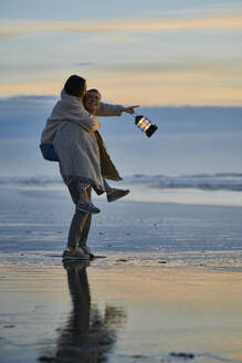 Junger Mann nimmt Frau mit Laterne am Strand huckepack - ANNF00904