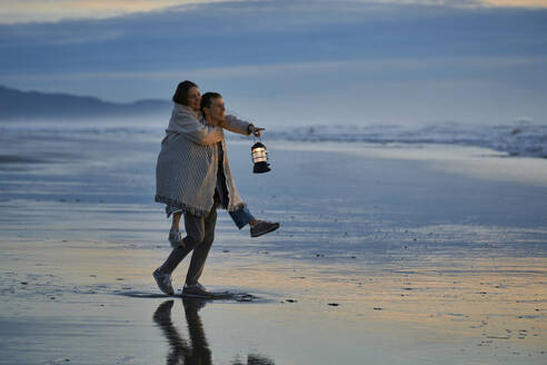 Junger Mann nimmt Frau mit Laterne am Strand bei Sonnenuntergang huckepack - ANNF00906
