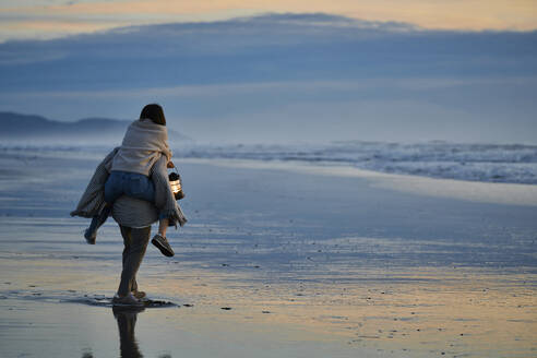Freund trägt Freundin huckepack und geht bei Sonnenuntergang am Strand spazieren - ANNF00908