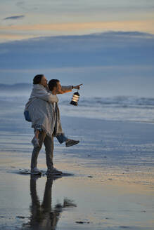 Mann nimmt Frau mit Laterne am Strand huckepack und zeigt auf das Meer - ANNF00911