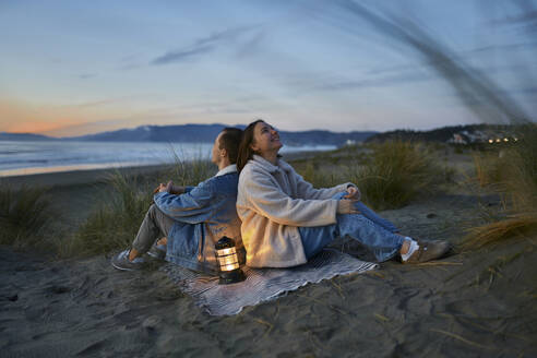 Nachdenkliches junges Paar mit Laterne auf Schal sitzend am Strand - ANNF00921