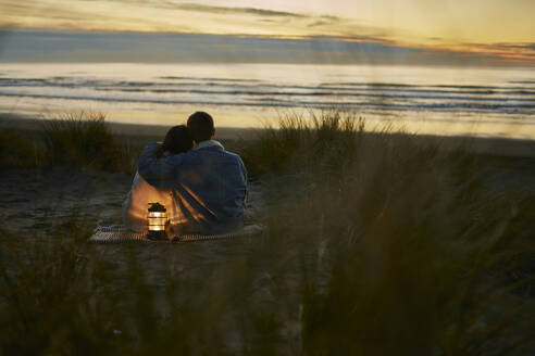 Junger Mann mit Arm um seine Freundin am Strand sitzend bei Sonnenuntergang - ANNF00929