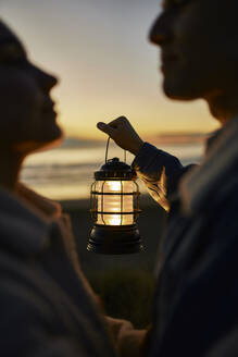 Young man holding lantern and looking at woman on beach - ANNF00933