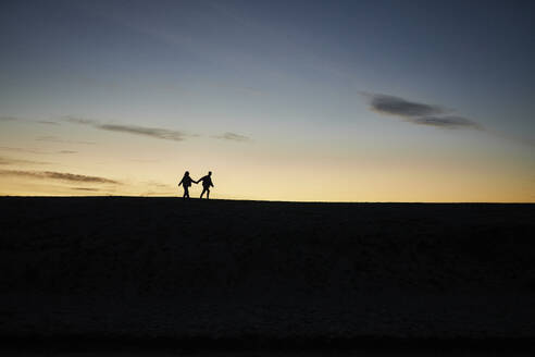 Silhouette eines jungen Paares, das sich an den Händen hält und am Strand spazieren geht - ANNF00939