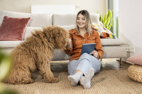 Smiling young woman petting dog and holding tablet PC at home - UUF31327