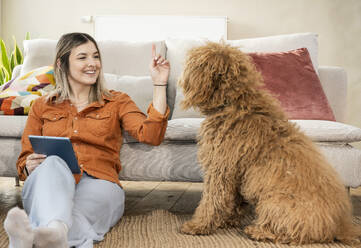 Smiling young woman gesturing and teaching dog at home - UUF31328