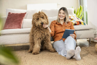 Happy beautiful woman holding tablet PC and stroking dog at home - UUF31329