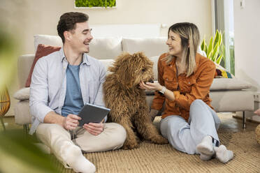 Happy couple sitting with dog and tablet PC at home - UUF31330
