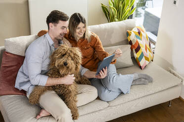 Glückliches Paar, das einen Tablet-PC benutzt und mit seinem Hund zu Hause auf dem Sofa sitzt - UUF31334