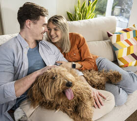 Glückliches junges Paar sitzt mit flauschigem Hund auf dem Sofa zu Hause - UUF31342