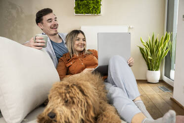 Glückliches Paar, das einen Laptop benutzt und in der Nähe eines Hundes zu Hause sitzt - UUF31345