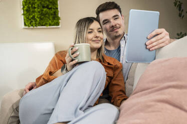 Lächelnde Frau hält Kaffeetasse und benutzt Tablet-PC mit Mann zu Hause - UUF31347