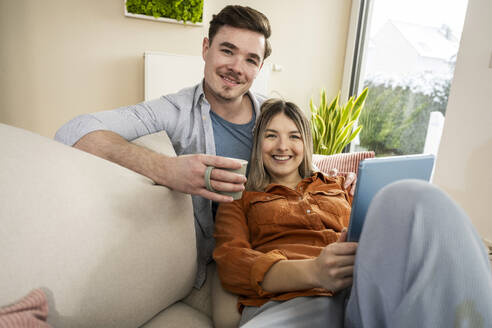 Glückliches junges Paar sitzt mit Tablet-PC und Kaffeetasse zu Hause - UUF31351