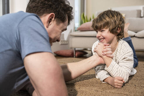 Happy son arm wrestling with father on carpet at home - UUF31390