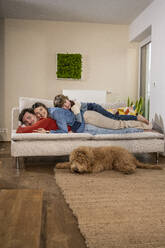 Happy father and mother lying on sofa near dog at home - UUF31394