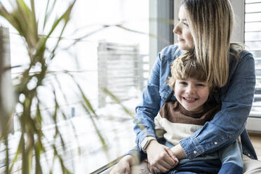 Glücklicher Sohn sitzt mit seiner Mutter am Fenster zu Hause - UUF31399