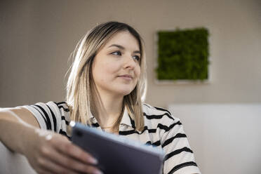 Lächelnde junge Frau, die mit einem Tablet-PC zu Hause sitzt - UUF31414