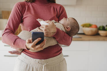 Mutter verwendet Smartphone und trägt süßes Baby in der Küche zu Hause - EBBF08726