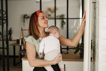 Smiling mother holding baby daughter and touching radiator at home - EBBF08728