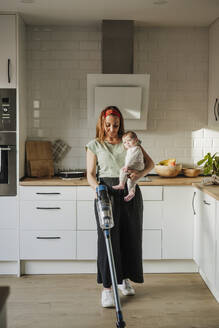 Frau trägt ihre kleine Tochter und putzt mit dem Staubsauger zu Hause - EBBF08736