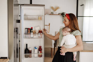 Lächelnde Frau, die ihre kleine Tochter trägt und die Kühlschranktür öffnet - EBBF08740