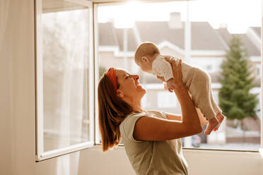 Happy woman playing with baby daughter near window - EBBF08741
