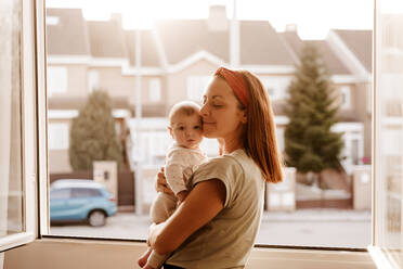 Mutter mit geschlossenen Augen umarmt Baby Tochter in der Nähe von Fenster - EBBF08743
