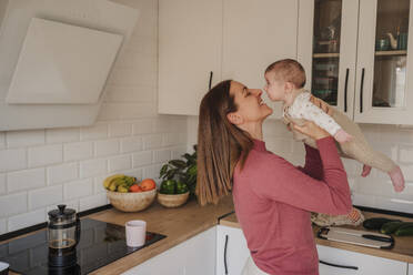 Smiling mother embracing and playing with baby daughter in kitchen - EBBF08749