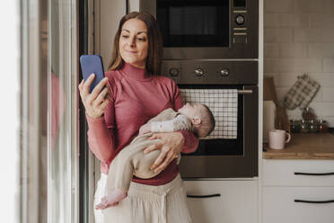 Smiling woman carrying baby girl and using smart phone in kitchen - EBBF08765