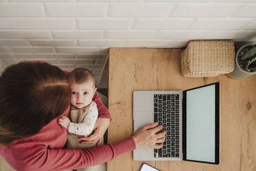 Freelancer mother typing on laptop and holding baby girl - EBBF08771