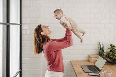 Happy freelancer mother playing with baby girl in kitchen at home - EBBF08775