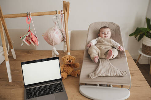 Cute baby girl sitting on bouncer chair near laptop at home - EBBF08785