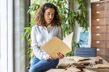 Geschäftsfrau liest Dokumente in der Nähe von hölzernen Proben auf dem Schreibtisch im Büro - DLTSF03778