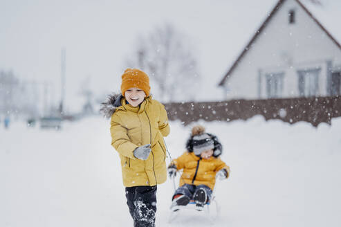 Happy boy pulling brother sitting on sled in winter - ANAF02717