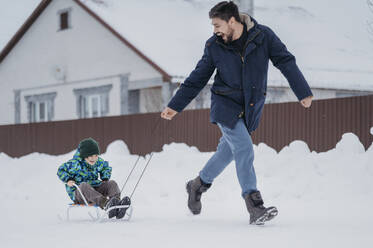 Vater zieht Sohn auf Schlitten sitzend im Winter - ANAF02720