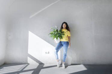 Lächelnde junge Frau, die eine Pflanze vor der Wand ihres neuen Hauses hält - AAZF01546