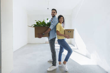 Glückliches Paar, das Kartons trägt und Rücken an Rücken im neuen Haus steht - AAZF01554
