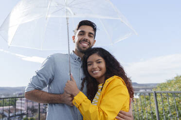 Glückliches Paar steht unter einem Regenschirm auf einem Balkon an einem sonnigen Tag - AAZF01561
