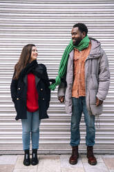 Lächelndes Paar mit gebundenen Schals vor einem Fensterladen stehend - AMWF01990