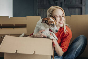 Lächelnde Frau umarmt Hund in der Nähe von Pappkartons zu Hause - VSNF01678