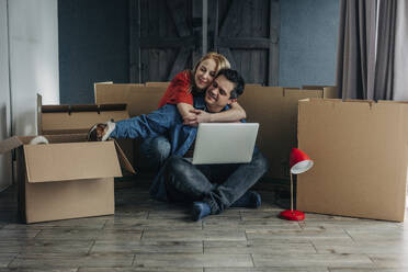 Happy woman embracing man sitting with laptop near cardboard boxes - VSNF01681