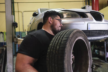 Mechaniker trägt Reifen in der Nähe des Autos in der Werkstatt - ASGF04921